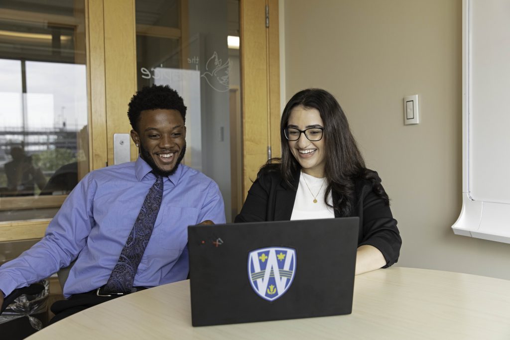 Two students, a male and a female, sit at a laptop which has the University of Windsor logo on it
