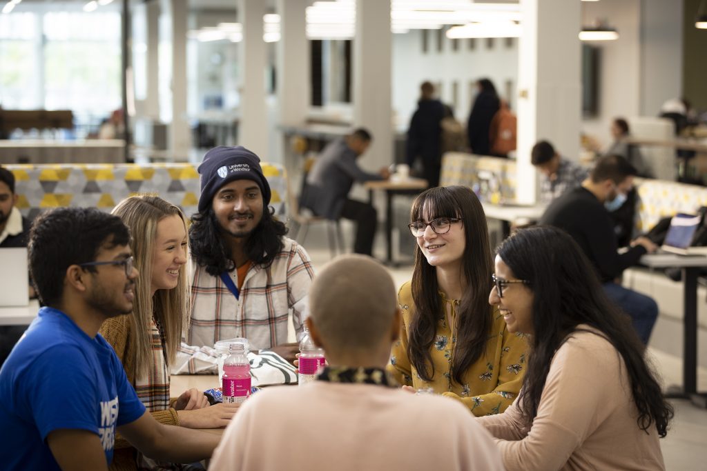 A group of smiling students sit at a table in the CAW Centre at the University of Windsor