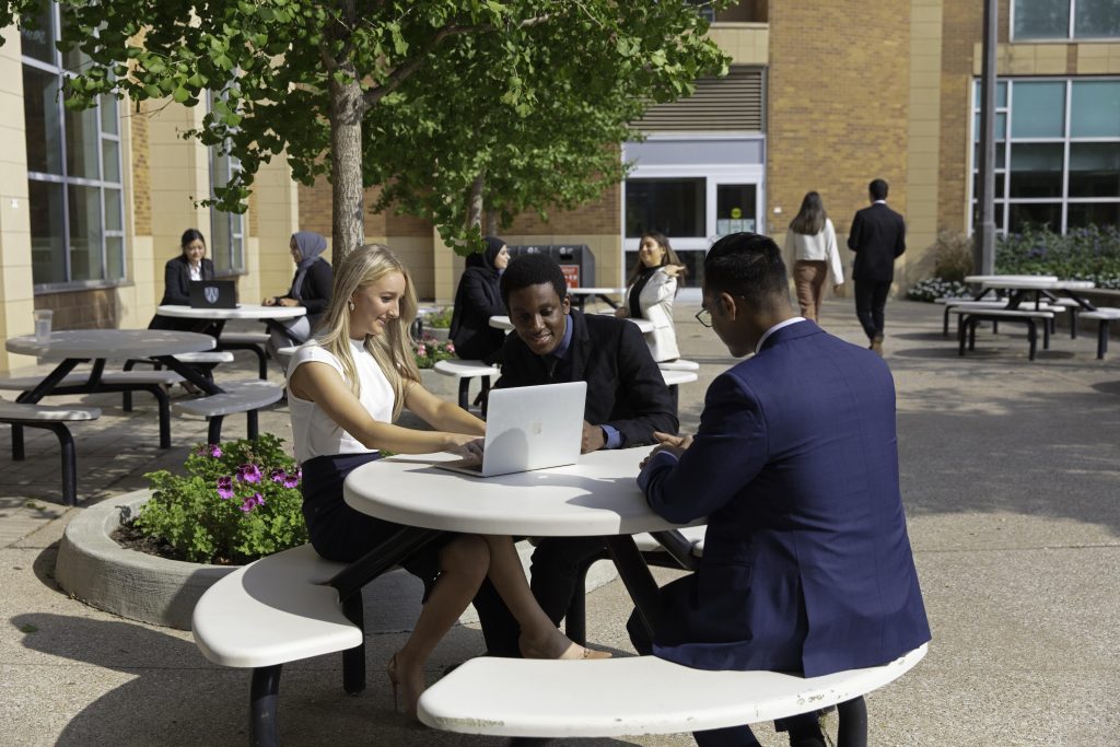 A group of students sit outside in business attire around a table looking at a laptop