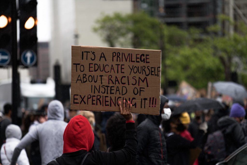 Photo of person holding a cardboard sign that says 'It's a privilege to educate yourself about racism instead of experiencing it'