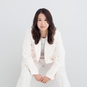 A photo of a Japanese woman in her 30s with medium length wavy brown hair. She is sitting on a white wooden box, in front of a white background. She is wearing a white pant suit. Under her blazer you can see a metallic silver crochet top layered over a white crop top. She is looking straight at the camera with a slight smile, and is leaning forward with he elbows on her knees and holding her hands in front. If you look carefully she's wearing silver rings.