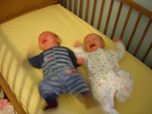 Two babies on their backs in a crib. Both are crying and kicking their legs.