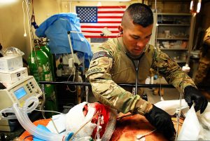 A patient with an endotracheal tube an severe facial trauma under a bandage is being assessed by a man in a US military uniform