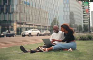 Two people sit on grass in the city smiling as they look on a laptop