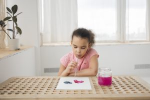 A little girl blows through a straw onto watercolour paint on a page to create art.