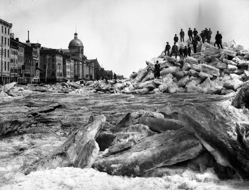 Black and white photograph taken from on the ice. Large pieces of protruding ice in the foreground and the rupture of the ice shove further back to the right, with several small and indistinguishable figures posed on top. The ice pushes against the stone buildings that run the length of the port on the left.