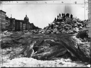 Black and white photograph taken from on the ice. Large pieces of protruding ice in the foreground and the rupture of the ice shove further back to the right, with several small and indistinguishable figures posed on top. The ice pushes against the stone buildings that run the length of the port on the left.