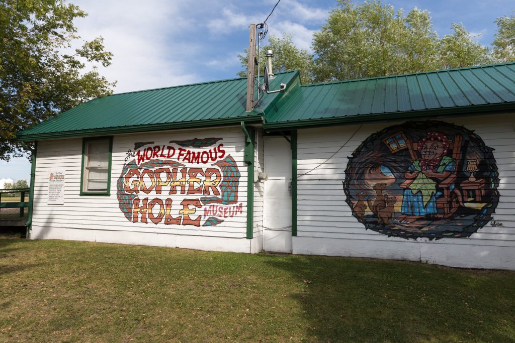 The exterior walls of the Gopher Museum featuring a textual mural which reads, “The World Famous Gopher Hole Museum,” in various decorative fonts and with an adjacent mural depicting a playful cartoon scene of a gopher hole, where a gopher dressed in matronly clothing knits in a rocking chair as three young gophers watch on.