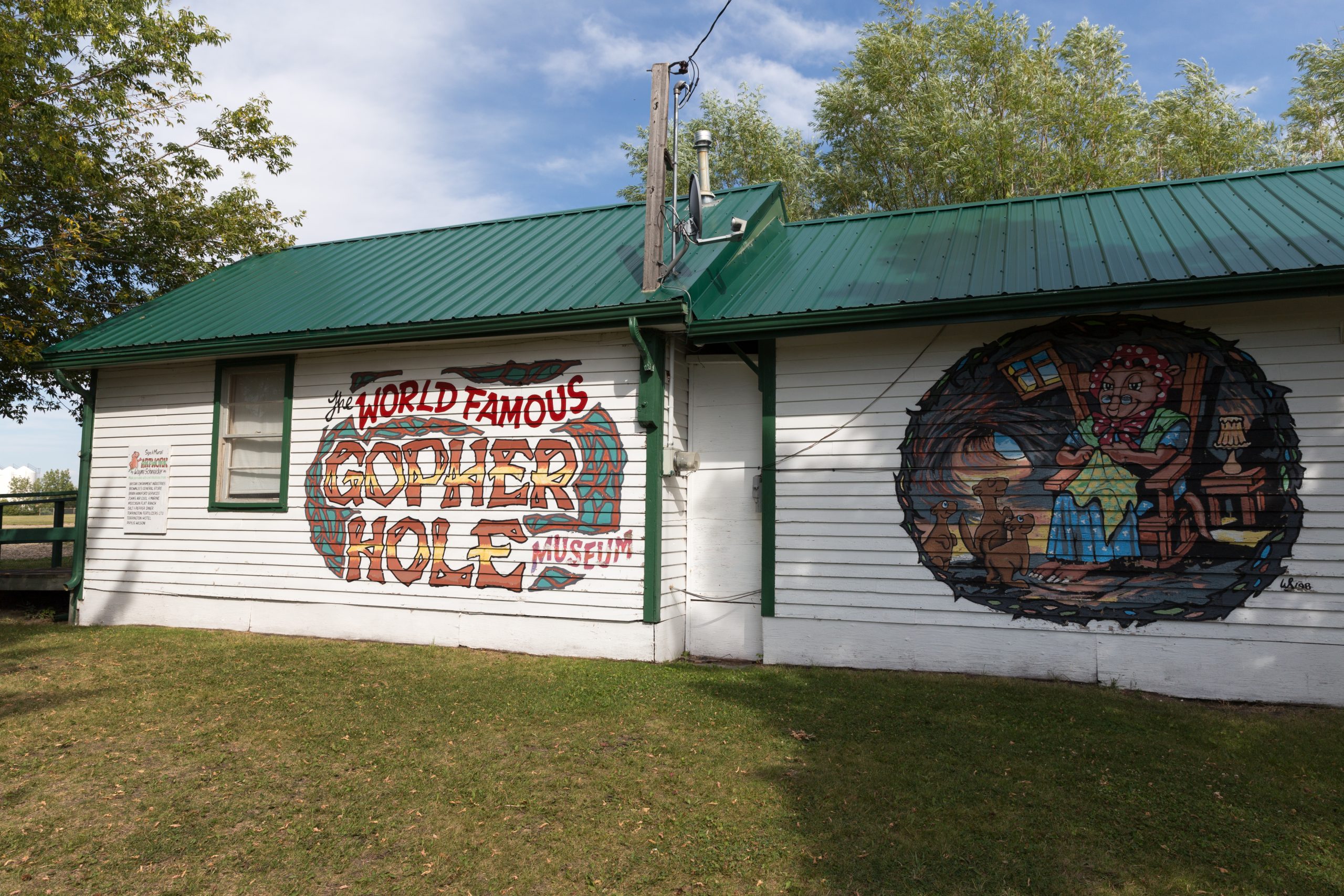 Exterior of a one-storey building with white siding and a green steel roof. The side of the building is hand-painted with the museum name and a colourful image of a gopher wearing a shawl, glasses and long dress, seated in a rocking chair near young gophers and knitting.