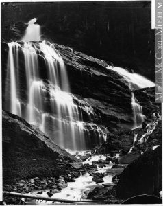 A stepped cliff dominates the black and white photograph, with a waterfall rushing from the top left, spreading out as it moves downwards until it flows swiftly in a rocky stream towards the viewer.