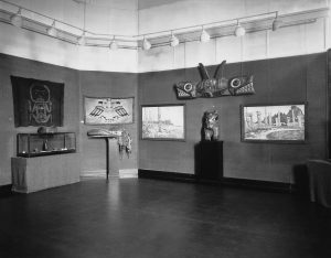 Black and white photograph of a room in a gallery. A series of four paintings—two of totem poles on beaches; two of stylized animal figures—hang from the walls. Above them hangs a painted wooden carving. A display case of smaller pieces is on the left, and two pieces—a mask and a sculpture—sit atop pedestals at the center and the right.