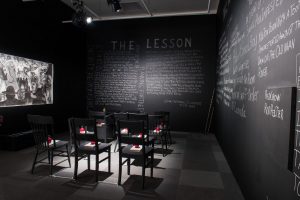 A dark photograph of a room with black chalkboard walls. The seven spot-lit black chairs have one white book and a red apple on each seat; the chalkboards they face and to their right are covered in writing in block letters; on the left-hand wall is a black and white image of many people with a small church at the centre. White ropes are attached to the chairs’ legs; the chairs cast stark shadows on the floor.