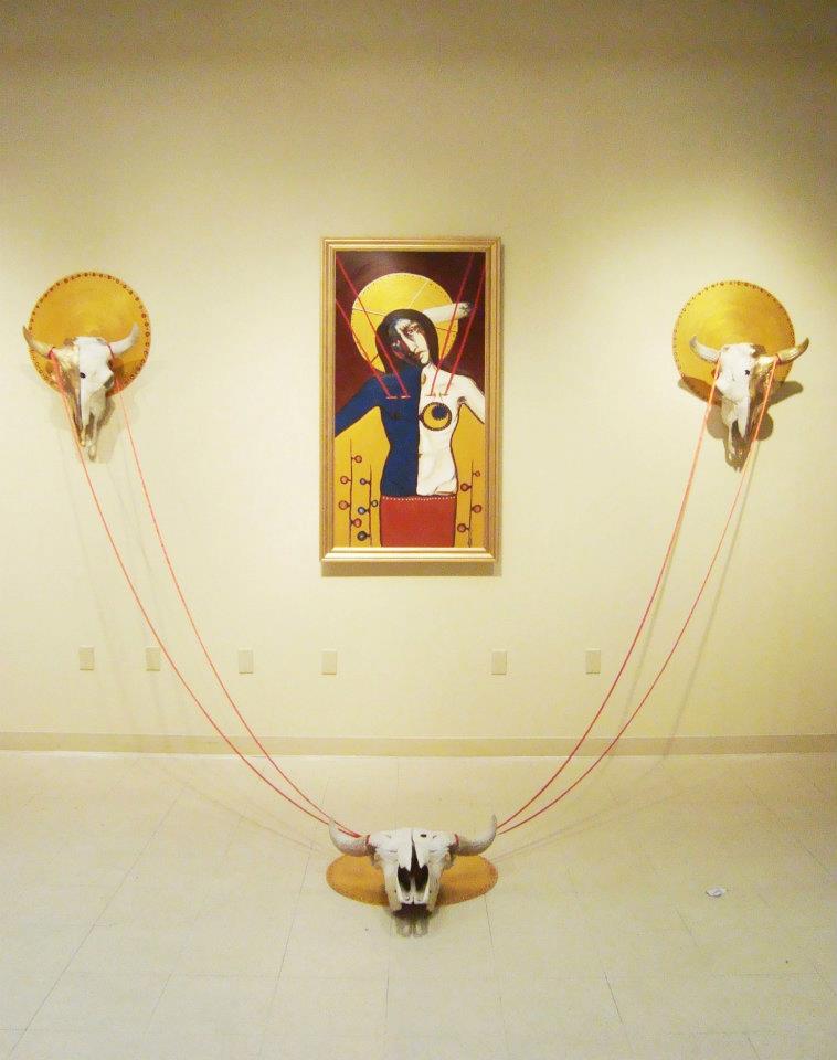 The installation with a painting depicting Jesus placed on the wall, and one buffalo skull on either side, connected by red string to a third buffalo skull placed on the ground, in front of the painting.
