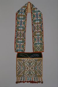 A flat, rectangular bag with a wide strap is covered in beading. Patterns are geometric and symmetrical in blue, gold, red, pink, white, green, and black. Red and gold tassels are at the end of each beaded fringe along the bottom of the bag.