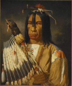 In this finished portrait, the features of the same man are smoother. He is dressed in elaborate Indigenous attire with adornments in his black hair and a staff decorated with a raven’s head and black and white feathers. He wears red paint on his face. The colours are richer and more varied; the background is shadowed ochre.