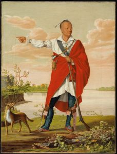 An Indigenous man stands facing the viewer in an open stance; his right arm is outstretched as he looks off to his left, in front of a lake and summer sky. He wears a silky white shirt, banded with silver cuffs and armbands, and over that, a red blanket draped over his left shoulder and around his torso. His head is mostly shaved with a feather attached to a lock of hair at the top. His left hand holds a long rifle with its butt on the ground and he has a pouch and pipe slung across his chest, beaded moccasins on his feet. A small brown and white dog looks up at him from the left.