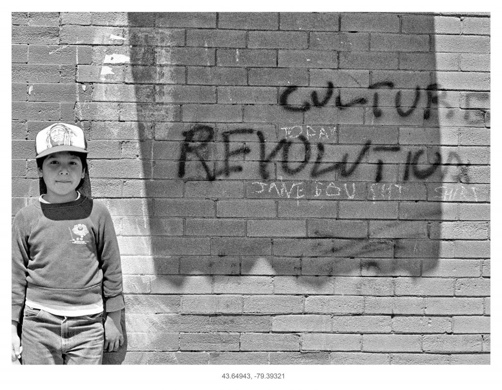 A boy in jeans, sweatshirt, and a ball cap with the image of an Indigenous chief on it stands against a brick wall. In the middle, an awning out of the shot casts a shadow onto the bricks. Next to the boy, the words “culture revolution” are spray-painted on the wall.