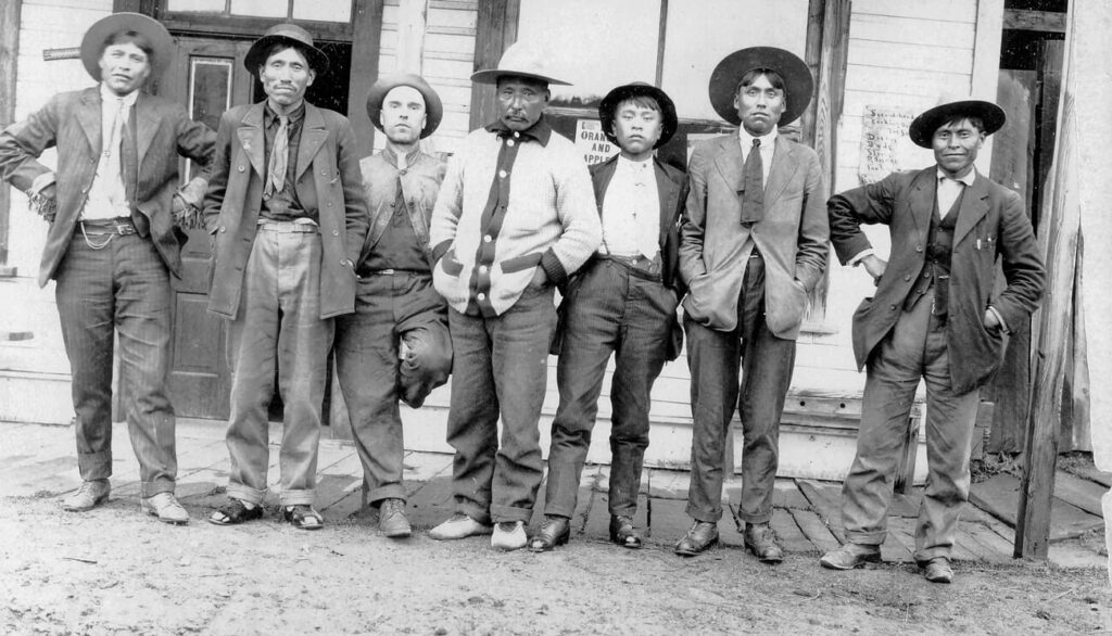 This black and white photograph shows seven men in front of a storefront. Dressed in shabby-looking shirts and jackets, hats and boots, only two wear ties or kerchiefs. All stare directly at the viewer.