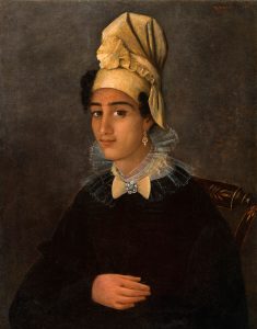 A young woman of colour sits in a chair and looks at the viewer in three-quarter profile. She wears a black dress with a white collar pinned with a jewelled brooch. A matching jewelled earring dangles from her left ear. Her dark hair is mostly contained in an elaborately folded golden headscarf. Her expression is contained but cheerful. Her right hand rests on her lap.