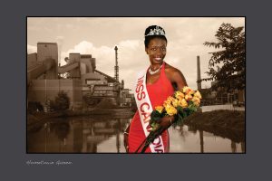 A black and white photograph of an industrial building in Hamilton, Ontario with the image of a smiling Black woman dressed as a beauty queen superimposed in colour. She wears a red dress, a necklace, a sash reading “Miss Canadiana,” and a tiara, and holds a bouquet of yellow roses.