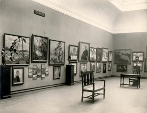 A black and white photograph of one long wall and a corner of a room in an art gallery with multiple pieces of art hanging almost to the floor. Three plinths hold statues at equidistant spaces against the walls. High on the wall above the artwork hangs a small plaque labelled “Canada”. A desk and two chairs stand in the middle of the room.