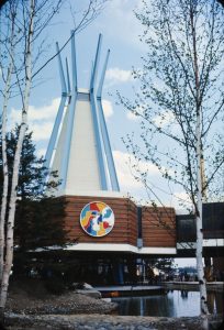In this colour photograph, a low wood-clad building with a roof tower resembling a tipi sits next to a pond with birch trees and rocks. On the side of the building is a large circular decoration in red, yellow, blue, and white.