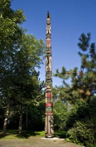 A large totem pole rises against a bright blue sky from a concrete sidewalk. Framed by trees in a parklike setting, its colours blend with its surroundings: brown, tan, dark green and black accentuated by red and bright green.