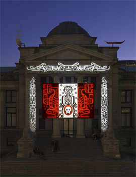 A colour photograph of the entrance façade of a neoclassical building against a dark blue-violet sky; the dim light obscures its architectural details. A design in bright white and red light is projected onto the entrance: white outlines of two totem poles are superimposed on the entrance columns and a similar but horizontal band appears just below the pediment. In the center is a design like the Canadian flag. The two sides display Indigenous Northwest designs in red on black backgrounds. The white center has a figure outlined in black with a skull on the lower portion, encircled in red.