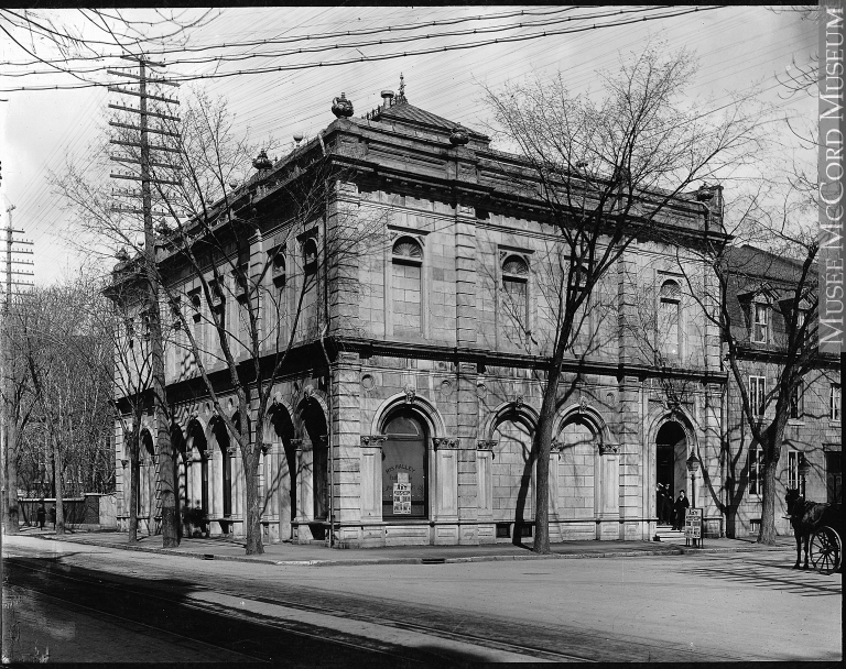 Black and white photograph of a neoclassical building on a city street corner, surrounded by bare trees and a lone telephone pole.