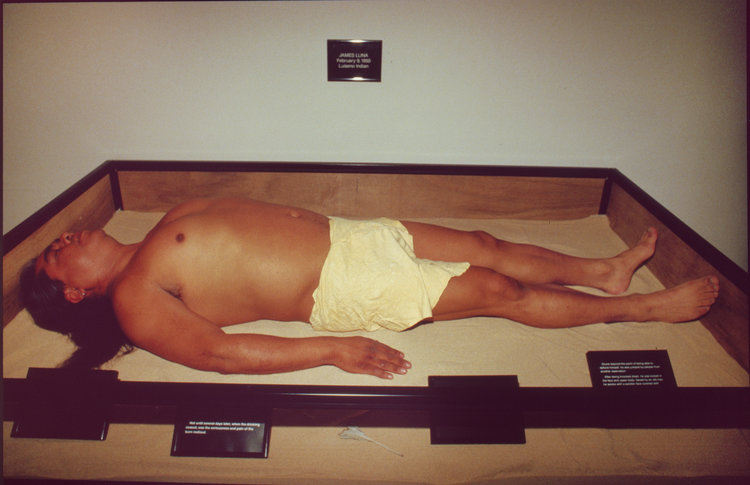 Colour photograph of an Indigenous man lying in a shallow box barely larger than him, wearing a white loincloth. His long hair is spread to the side. The box is plain wood with its edges painted black; its bottom is lined with a cream-coloured fabric.