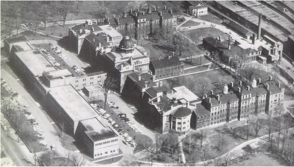 The photograph is an aerial view of a sprawling institutional complex with multiple buildings that appear to be added at different times. The u-shaped classicized building dominating most of the landscape in which it sits is composed of a central flank topped with a dome and connected to two parallel wings stretching southward. The wings appear to be composed of several 19th century townhouses of varying heights. Directly in front of the domed central flank sits a 1950s institutional building, which hides the older complex from view of the street onto which it fronts.