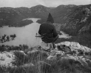Photograph of Franklin Carmichael sitting atop a vantage point, sketching the view of Grace Lake and the surrounding La Cloche mountains.
