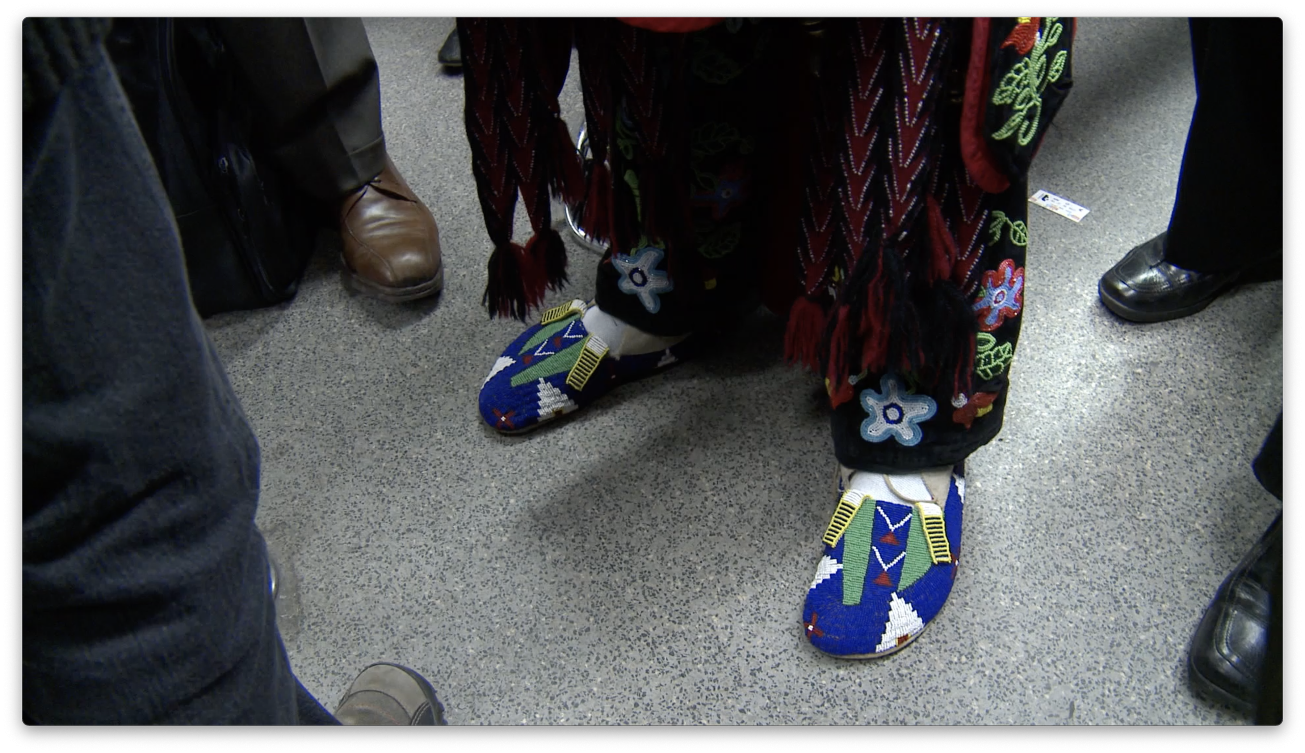 Colour film still of a man dressed in full beaded Regalia. He is wearing a feather headdress and holding Eagle feathers, porcupine quills, and a traditional war club. The man is at the bottom of the stairs in a Paris metro station, beside barriers for ticketed entry and exit. Behind him on the stairs are passengers including a man in a suit who is holding a cell phone, taking a picture.
