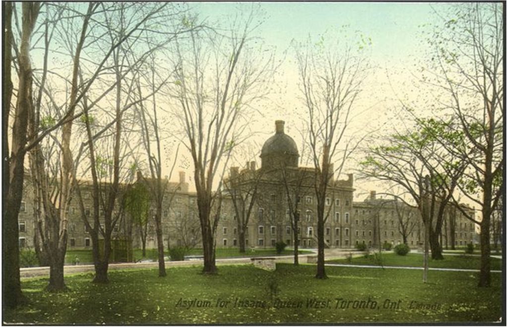 A colourized postcard showing the façade of a sprawling 19th century institution with a central dome set within a grassy landscape. The view is diagonally angled towards the building, which is partially obscured by trees in various stages of early spring leaf out. Paths lead through the manicured green landscape. At the bottom-right of the postcard, the following is printed in black stamped text: Asylum for Insane. Queen West, Toronto, Ontario, Canada.