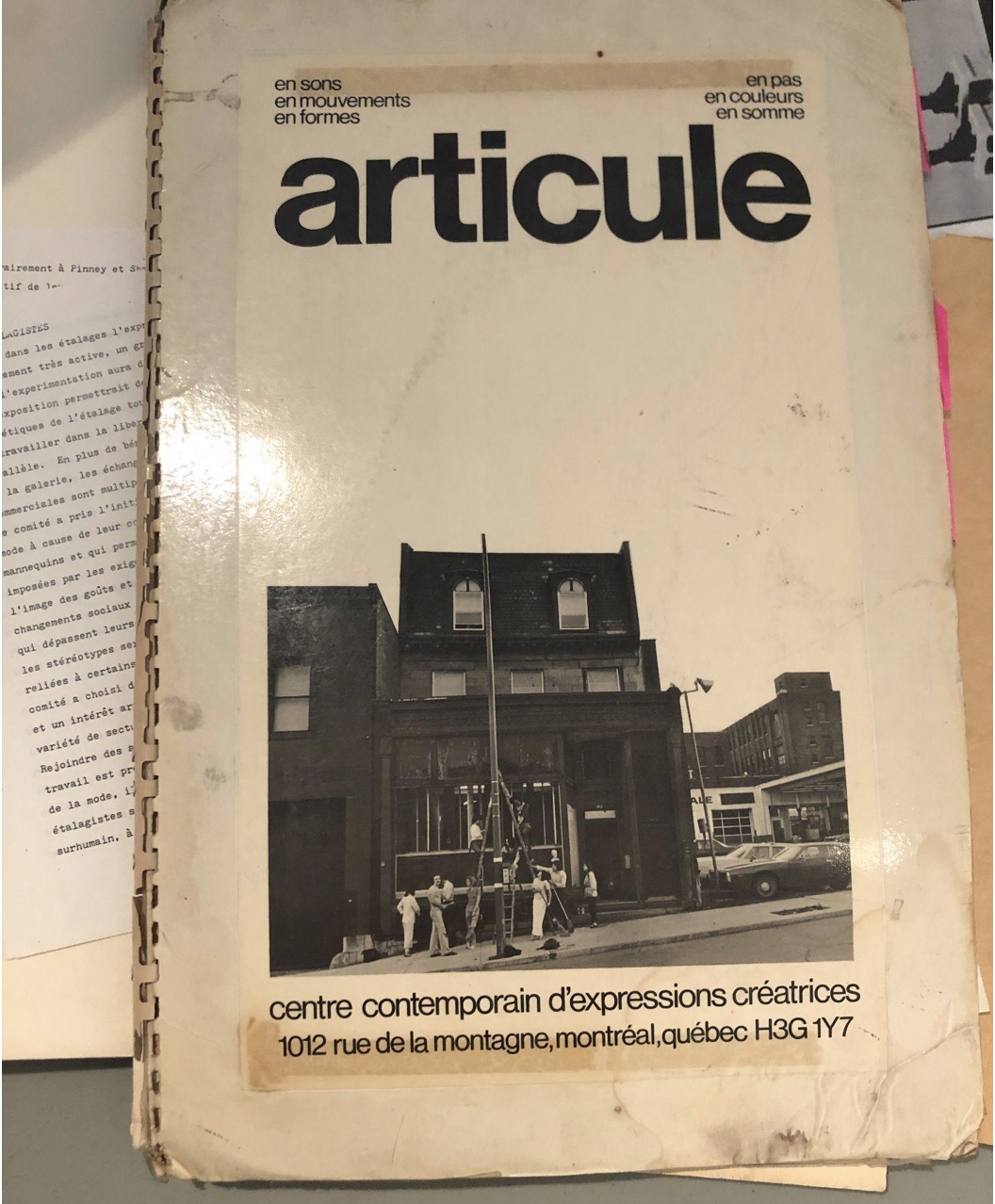 Image of archival documents featuring a catalogue displaying a photograph of the articule original storefront circa 1980 on de la Montagne street in Montreal. Several members stand in front of an erect ladder installing work in front of the large gallery window.