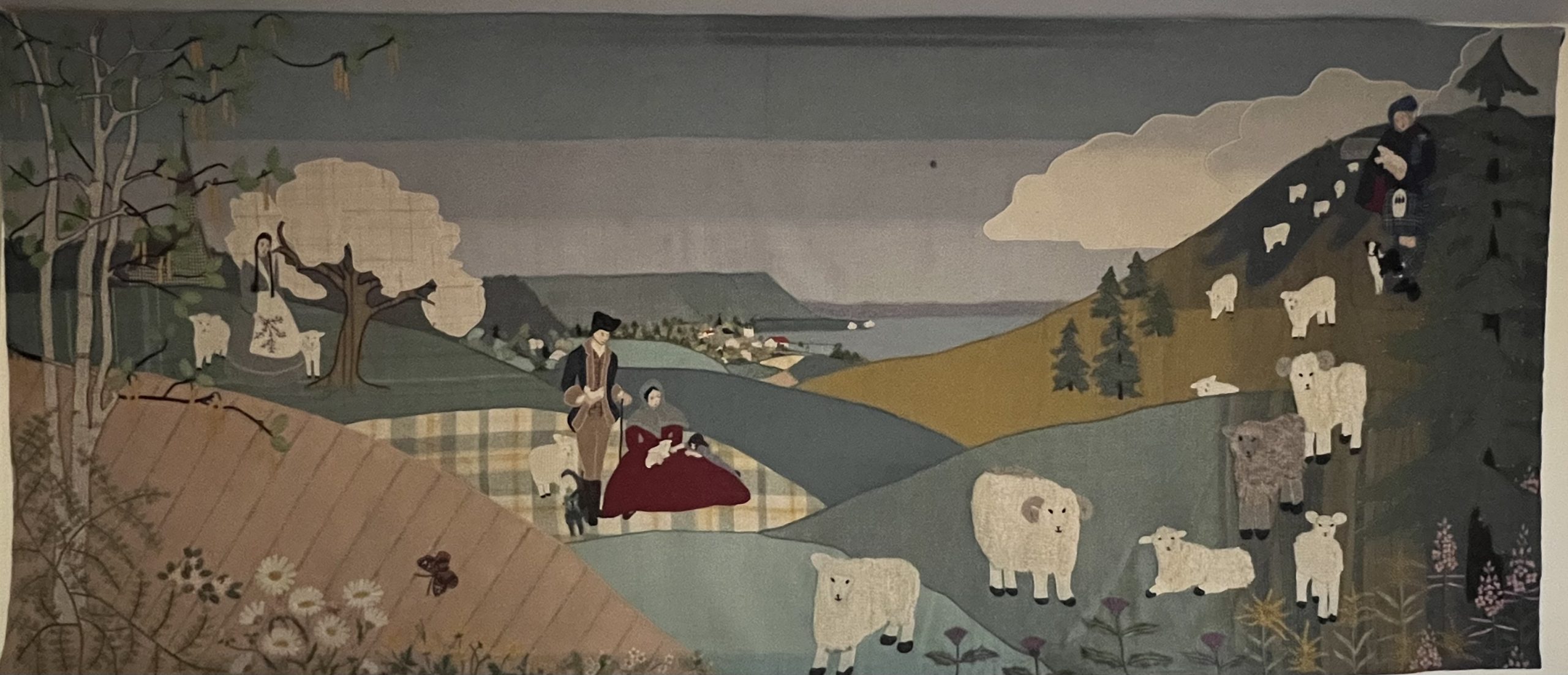 A large rectangular mural composed of appliquéd wool sections in predominantly muted colours. On the far left, a white girl dressed in Acadian folk costume tends sheep beneath an apple tree. In the centre, a white family dressed as United Empire Loyalists are resting on a hill. On the far right, a white man in a blue and green great kilt herds sheep.