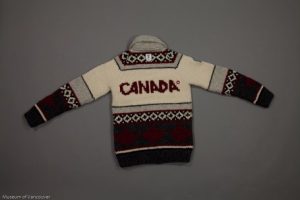 On a gray background, a knitted sweater is spread out flat and face-down, showing the word “CANADA” across the back. Coloured bands are in warm cream, grays, and black with a red and white Olympic patch on the right sleeve and a band of dark red maple leaves around the bottom.