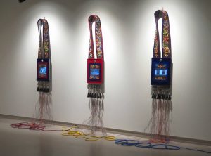 Three bandolier bags hang from a light gray wall. Each bag has a screen displaying decorations attached to the front of it in lieu of traditional beadwork, but colourful beadwork decorates each strap and tassels hang below each bag. Red, yellow, and blue electrical cords connected to each bag lie on the floor below them.