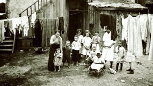 A white woman, a Black woman and eight children stand in a group and look at the photographer. Two babies are in strollers. Another child’s legs peek out from behind a clothesline. They stand on a litter-strewn patch of dirt surrounded by ramshackle buildings and clotheslines full of laundry.