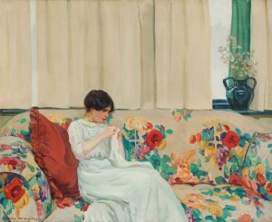 A dark-haired white woman in a pale green dress sits on a large sofa, leaning against a red throw pillow, sewing a piece of white fabric. The sofa is upholstered in a beige fabric with large red, yellow, green, purple and pink flowers. The beige drapes in the window behind are pulled shut except for a small section on the right side where a dark green vase with white flowers sits. A swath of green fabric hangs directly behind the vase.