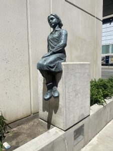 A bronze sculpture of a young girl dressed in European clothing on the east side of the work, sitting on a stone plinth with her hand resting on her lap.