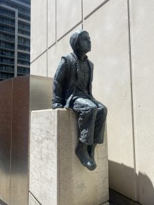 A bronze sculpture of a young boy on the west side of the work, dressed in traditional Chinese clothing, sitting on a stone plinth with his hands placed beside him on the stone.