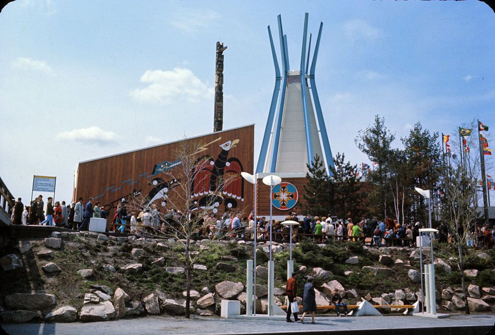 Color photograph of the exterior of the Indians of Canada Pavilion at Expo 67. The pavilion is in the form of an abstracted tipi, with bright blue steel beams serving as “poles” and white vinyl cladding. Other elements of the pavilion are clad in cedar boards, with artwork hung or painted directly on the surface. A totem pole emerges from behind part of the pavilion and a crowd of visitors is lined up in front.
