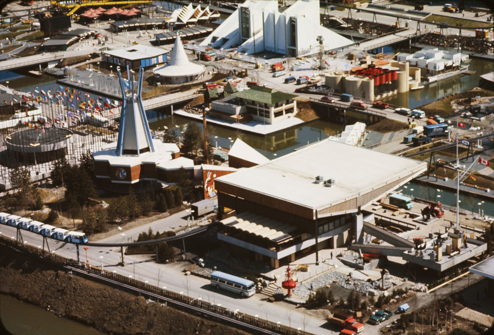 An aerial view of part of the Expo 67 site on the southern end of Île Notre-Dame, Montreal. The Indians of Canada Pavilion is towards the left of the image. The United Nations Pavilion, a short, cylindrical building surrounded by flags is to its left. The Atlantic Provinces Pavilion, a large building with rectilinear forms of different heights, is to its right. The Expo monorail is in the foreground, approaching a curved part of the track leading into the Atlantic Provinces Pavilion. Canals, bridges, and other pavilions are in the background.