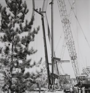 A black-and-white photograph of the Indians of Canada Pavilion under construction. A crane on the right is placing a steel beam, with three others already in place. Two workers stand at the top of the construction, and three others are working with the beam at the bottom. Several other figures in white construction hats stand near the crane on the bottom right. On the left, a pine tree stands in the foreground.