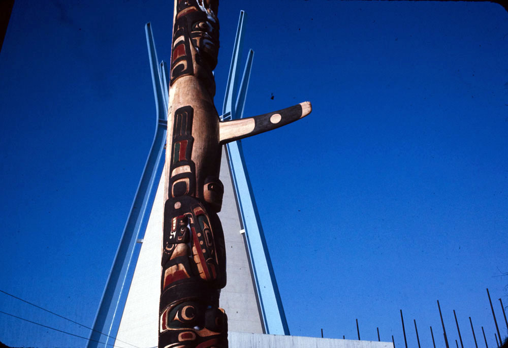 In the foreground, a crowd of spectators watches a performance of the Raven Dance by Harry Hunt in front of the Indians of Canada Pavilion. Behind them, to the left, is part of the totem pole carved by Henry and Tony. To the left, hung on part of the pavilion, is Alex Janvier’s The Unpredictable East (Beaver Crossing, Indian Colours), a circular, abstract painting with converging forms in red, yellow, white, blue, and dark blue. The windows of the pavilion behind are covered in clear plastic sheeting.