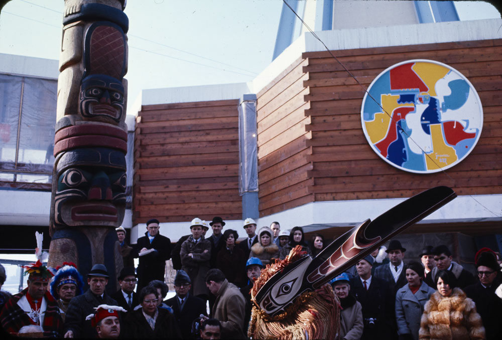 Harry Hunt performs the Raven Dance for a crowd of spectators in front of the Indians of Canada Pavilion. Behind the crowd, to the left, is part of the totem pole carved by Henry and Tony Hunt. To the right, hung on part of the pavilion, is Alex Janvier’s The Unpredictable East (Beaver Crossing, Indian Colours), a circular, abstract painting with converging forms in red, yellow, white, blue, and dark blue.