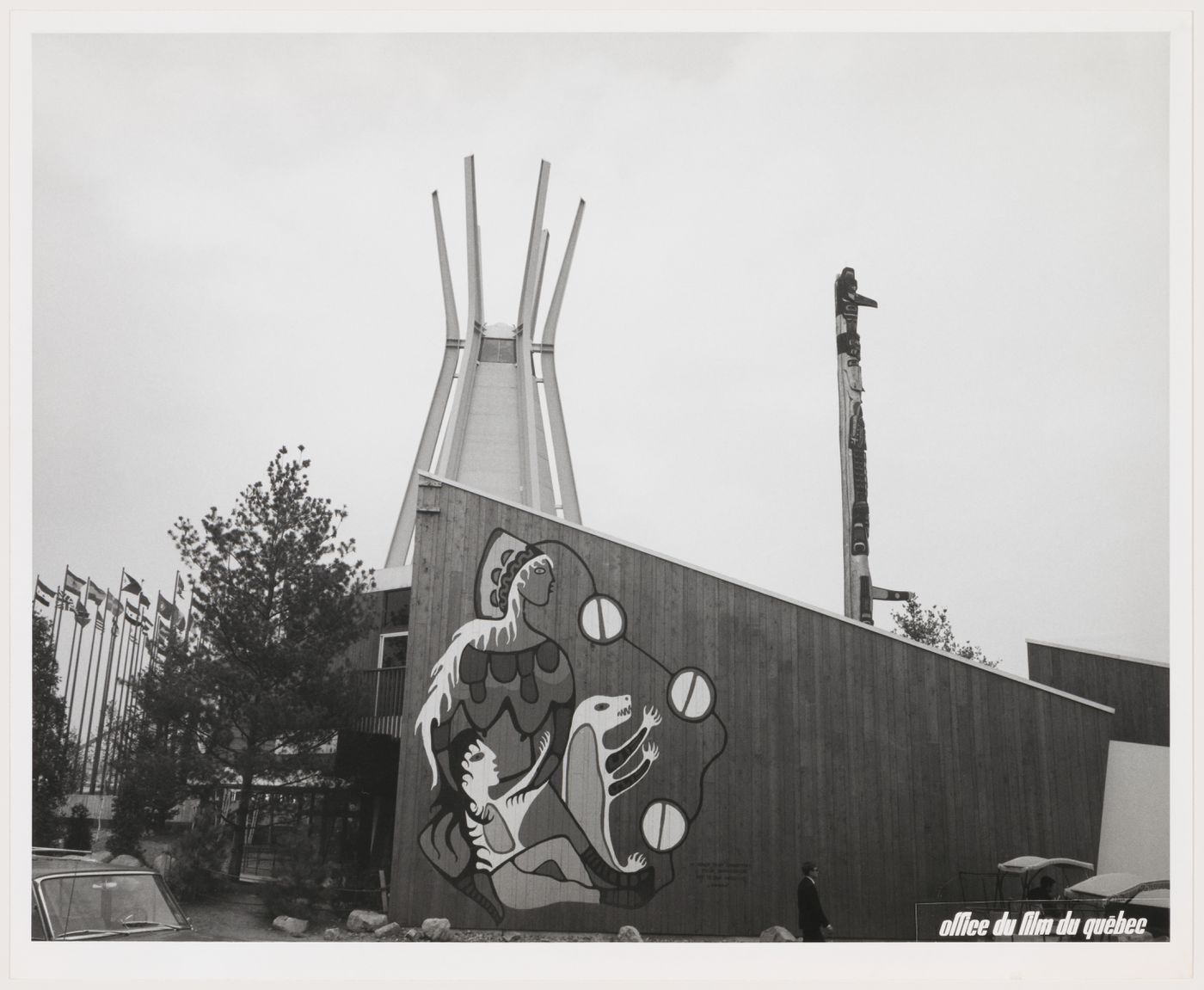 A black-and-white photograph of Anishinaabe artist Norval Morrisseau’s mural at the Indians of Canada Pavilion. The mural, painted directly on the vertical cedar boards of the building, depicts the Earth Mother with long, white hair, a head covering, and exposed breasts, along with a child with long black hair, and a white and yellow bear cub. A series of circular forms linked with black lines connects the figures to the ground plane. The “tipi” and totem pole are visible in the background.
