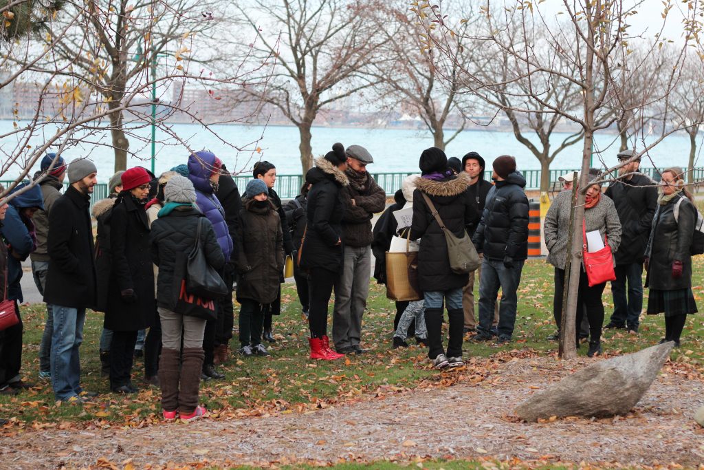 A colour photograph documenting a performative tour. A crowd wearing winter clothing gathers in an orchard next to a river. Some people carry sheets of paper, and a central figure with their back to the camera seems to be a focal point for the group. The group stands on orange and brown leaves and green grass next to the pathway.