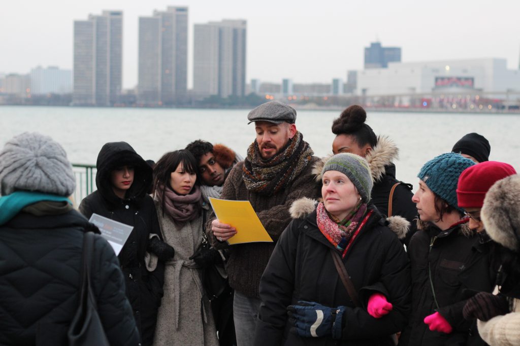 A colour photograph documenting a performative tour. A tour group dressed for winter weather gathers around two central figures standing on either side of a commemorative bronze plaque, the heading of which reads “THE FRANÇOIS BABY HOUSE LA MAISON BABY.” A brick building is in the background. One figure carries a white binder, and the other figure is speaking to the group.